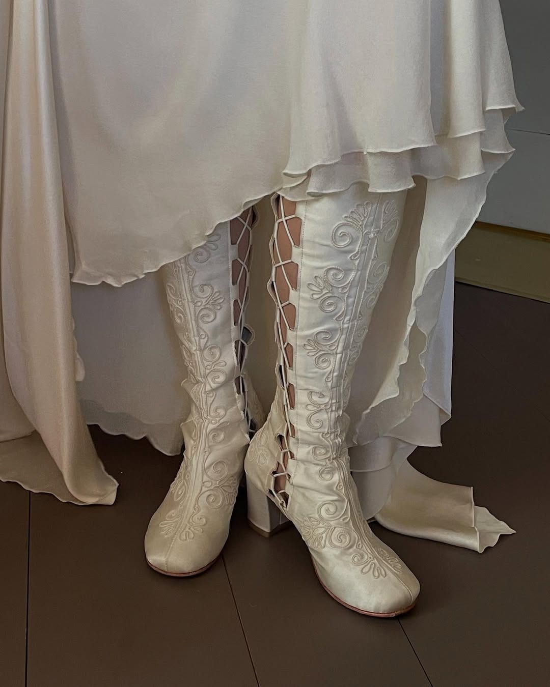 White embroidered knee-high boots worn with a white dress on a wooden floor.